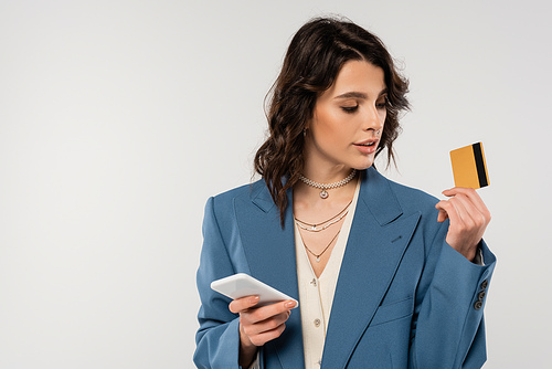 stylish brunette woman holding smartphone and looking at credit card isolated on grey
