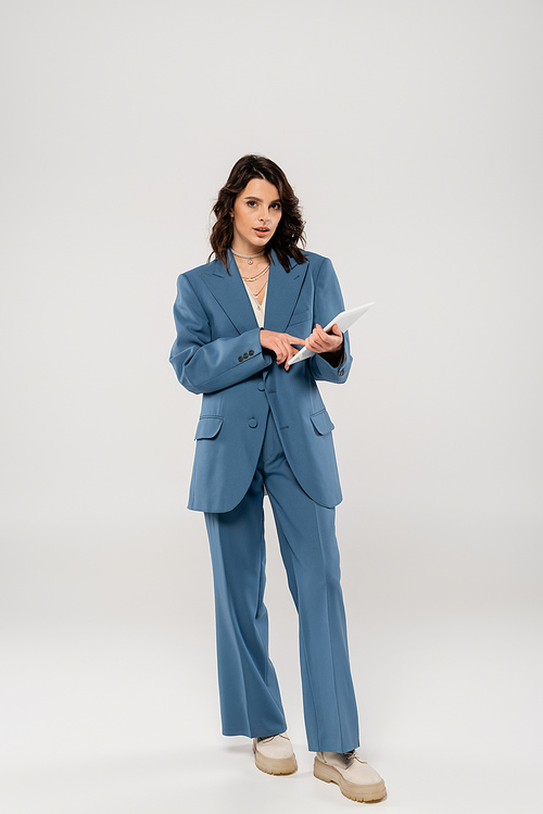full length of trendy woman in blue suit using digital tablet and looking at camera on grey background