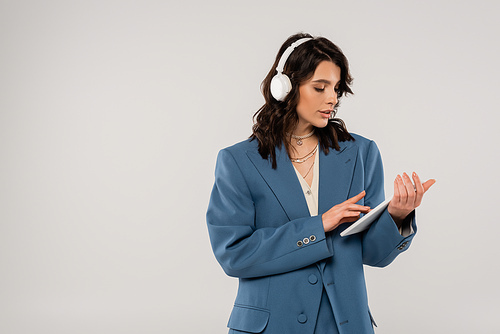 brunette woman in wireless headphones and blue blazer using digital tablet isolated on grey