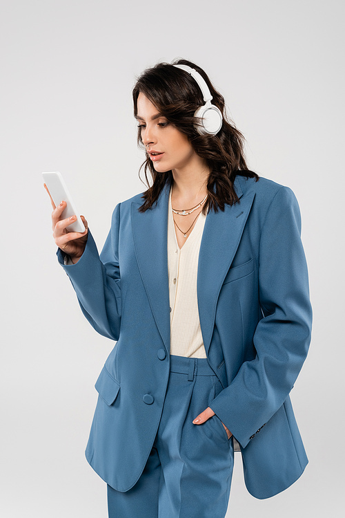 trendy woman in wireless headphones standing with hand in pocket of blue pants and looking at smartphone isolated on grey