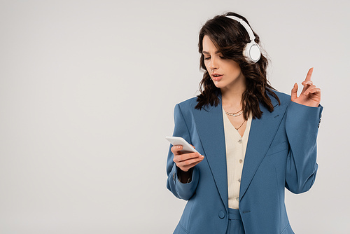 trendy brunette woman in wireless headphones looking at smartphone and pointing with finger isolated on grey