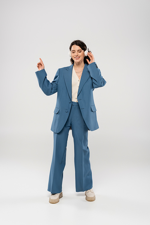 full length of joyful woman in blue suit listening music in wireless headphones on grey background