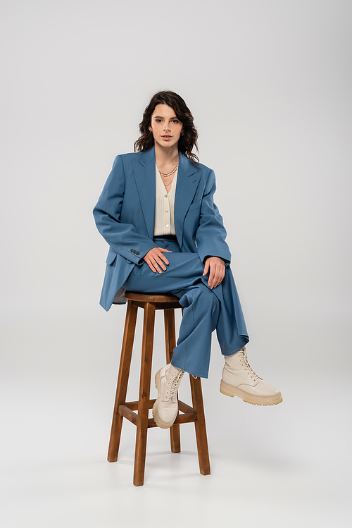 full length of brunette woman in blue suit and white boots sitting on wooden stool on grey background