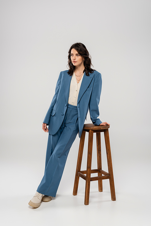 full length of brunette woman in blue suit posing near wooden stool on grey background