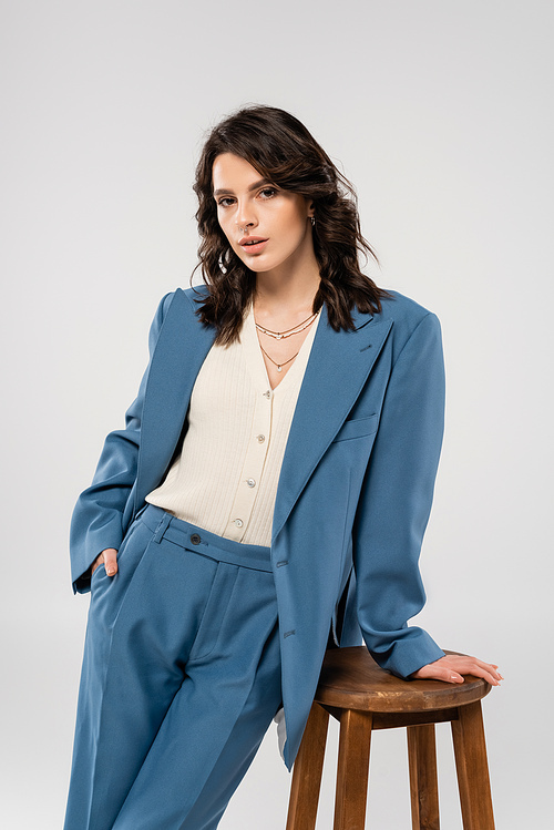 trendy woman in white jumper and blue suit posing with hand in pocket near wooden stool isolated on grey