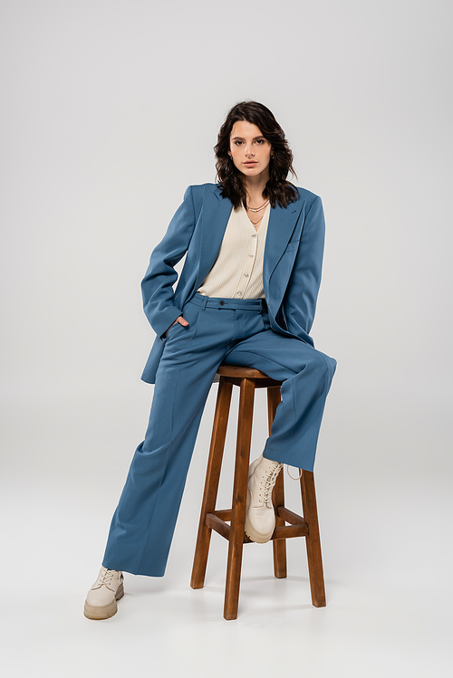 full length of young woman in blue and stylish suit holding hand in pocket while posing on stool on grey background