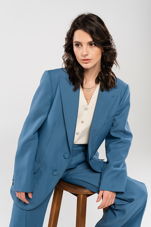 trendy brunette woman in blue blazer sitting on wooden stool and looking away isolated on grey