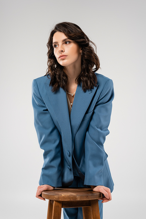 young and stylish woman in blue jacket looking away near wooden stool isolated on grey