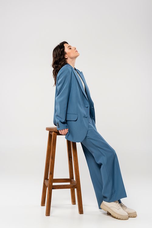 side view of brunette woman in blue suit and boots leaning on wooden stool on grey background