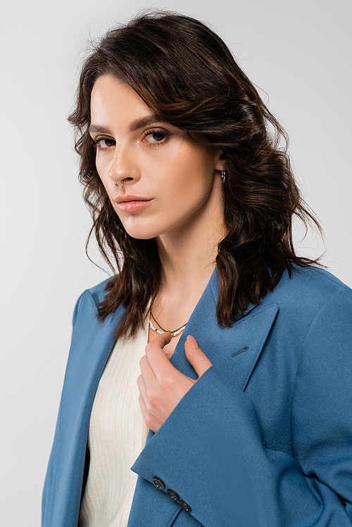 portrait of young brunette woman in blue stylish jacket looking at camera isolated on grey