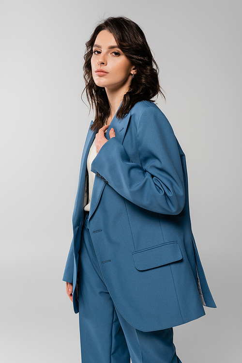 trendy brunette woman in oversize blazer looking at camera isolated on grey