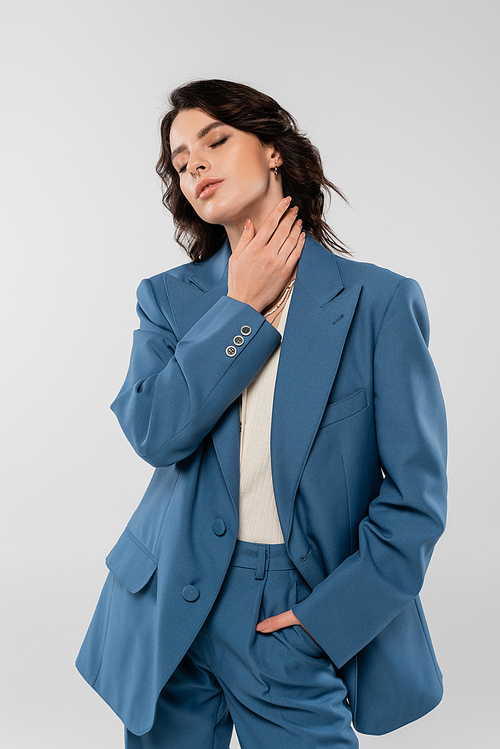 sensual brunette woman in blue suit holding hand in pocket and touching neck while standing with closed eyes isolated on grey