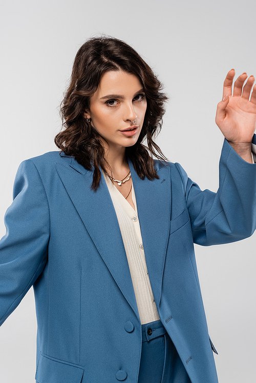 brunette woman in blue stylish jacket posing with outstretched hand isolated on grey