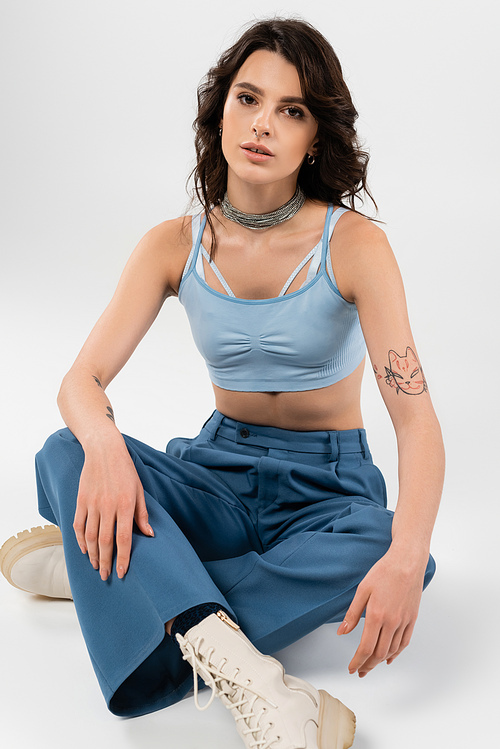 tattoed woman in blue crop top and pants with leather boots sitting with crossed legs on grey background