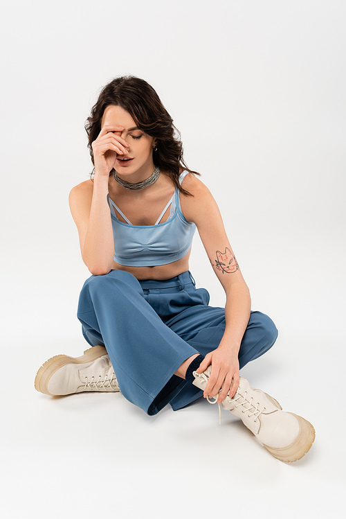 brunette woman in blue crop top and pants with laced boots sitting with crossed legs and obscuring face with hand on grey