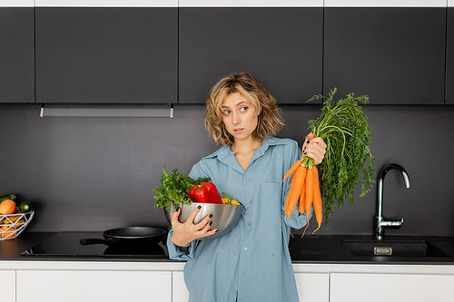 confused young woman holding bowl with vegetables and fresh carrots in kitchen