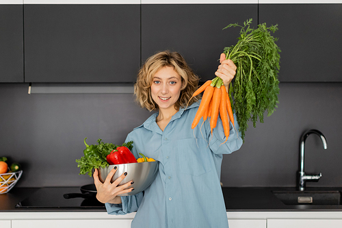 cheerful young woman holding bowl with vegetables and fresh carrots in kitchen
