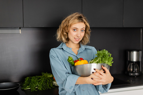 cheerful young woman holding bowl with fresh vegetables in kitchen