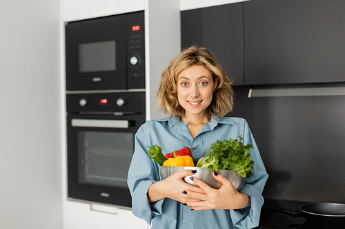 positive young woman holding bowl with fresh vegetables in kitchen