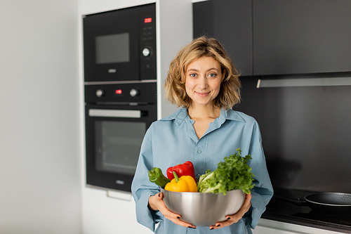 pleased young woman holding bowl with fresh vegetables in kitchen
