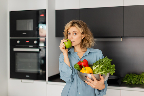 blonde young woman holding bowl with fresh vegetables and green apple in kitchen