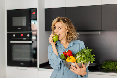 blonde woman biting apple and holding bowl with fresh vegetables in kitchen