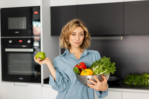 young woman with wavy hair holding bowl with fresh vegetables and apple in kitchen