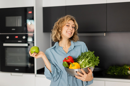 happy young woman with wavy hair holding bowl with fresh vegetables and apple in kitchen