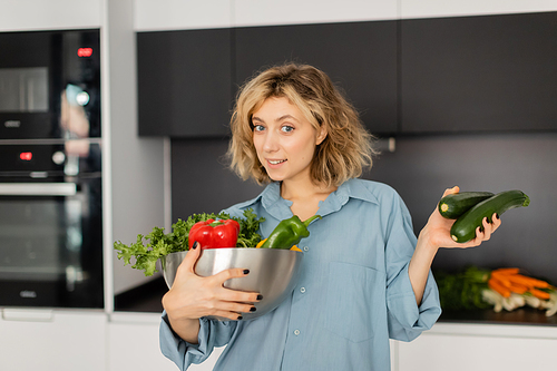 happy young woman with wavy hair holding bowl with fresh vegetables in kitchen