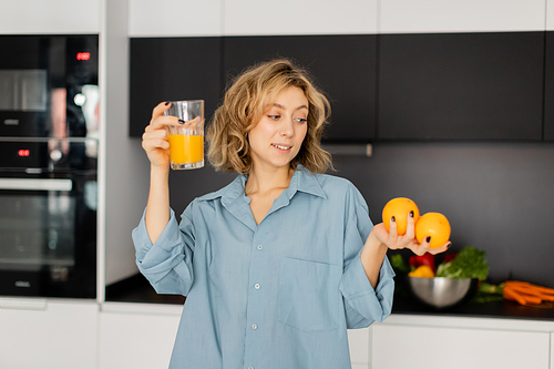 pleased young woman with wavy hair holding fresh oranges and glass with juice in kitchen