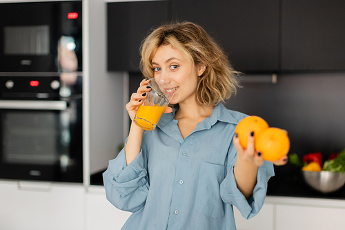 happy young woman with wavy hair holding fresh oranges and drinking juice in kitchen