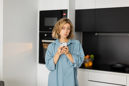 pensive young woman with wavy hair holding smartphone in kitchen
