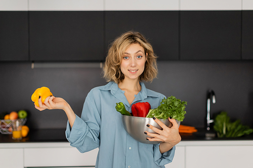 cheerful young woman with wavy hair holding bowl with organic vegetables in kitchen