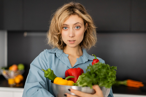 blonde young woman with wavy hair holding bowl with organic vegetables in kitchen