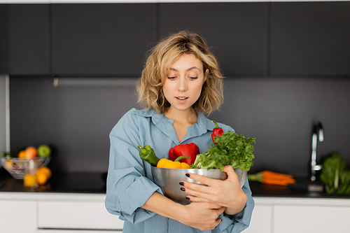 pretty young woman with wavy hair holding bowl with organic vegetables in kitchen