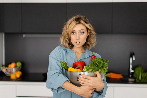 surprised and blonde woman with wavy hair holding bowl with organic vegetables in kitchen