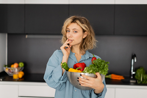 blonde young woman with wavy hair holding bowl with organic vegetables and looking at camera