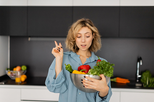 blonde young woman with wavy hair holding bowl with organic vegetables and showing idea sign