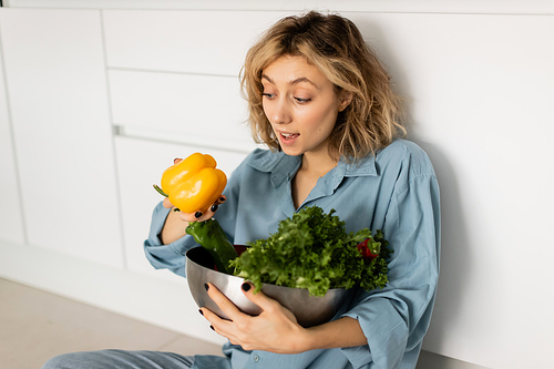 amazed blonde young woman with wavy hair holding bowl with tasty vegetables in kitchen