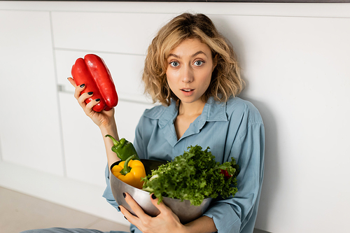 shocked young woman with wavy hair holding bowl with organic vegetables in kitchen