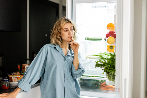 pensive young woman looking into open refrigerator in kitchen