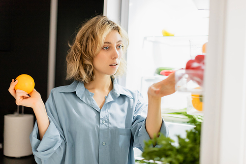 thoughtful young woman holding orange and looking at refrigerator in kitchen