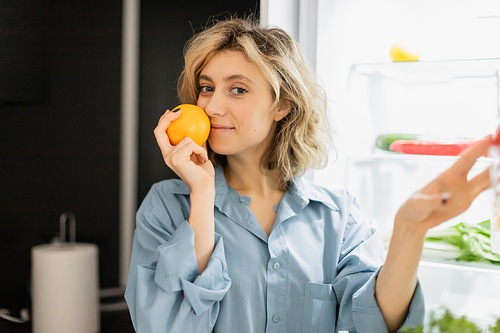 happy young woman smelling orange near refrigerator in kitchen