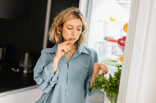 pensive young woman looking at greenery in open refrigerator