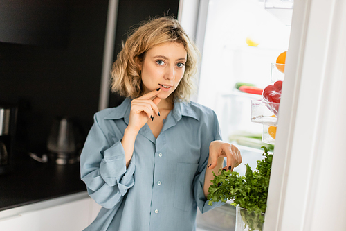 pensive young woman looking at camera near open refrigerator in kitchen