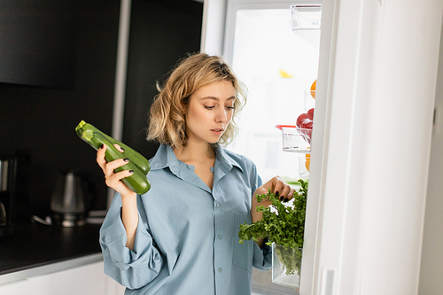 blonde young woman in blue shirt looking at greenery while holding zucchini near open refrigerator in kitchen