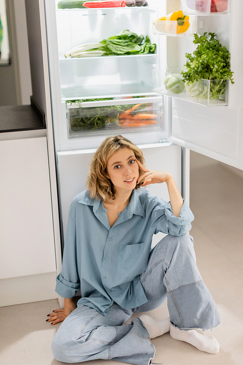 happy young woman sitting near opened refrigerator with fresh products in kitchen