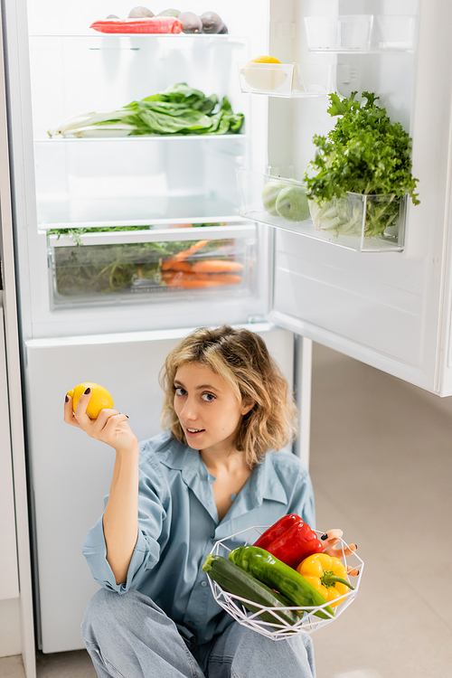 young woman sitting near opened refrigerator and holding bowl with fresh vegetables in kitchen