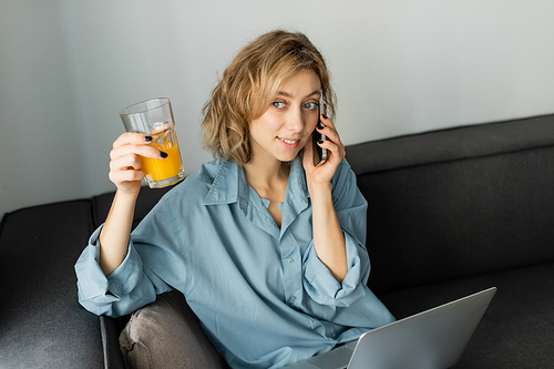 cheerful freelancer with wavy hair holding glass of orange juice while talking on smartphone near laptop