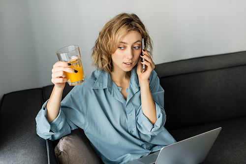 high angle view of young freelancer with wavy hair holding glass of orange juice while talking on smartphone near laptop